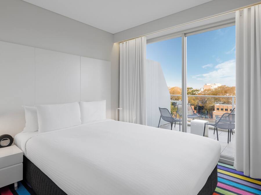 Una habitación de hotel moderna con una cama grande, una mesita de noche, un escritorio y un balcón con sillas.
