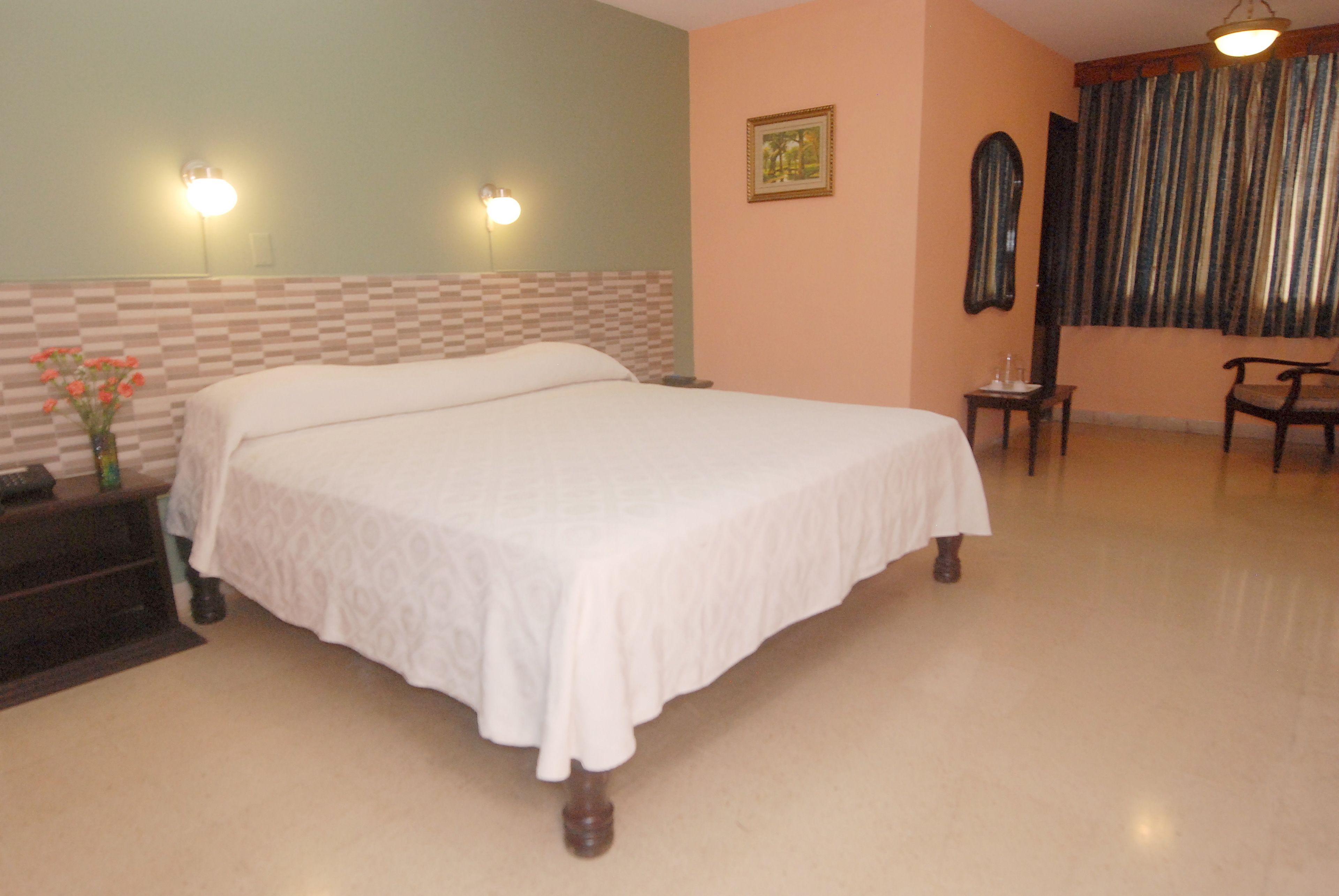 A hotel room with a large bed, two wall-mounted lights, a small table, a chair, and a window with curtains.