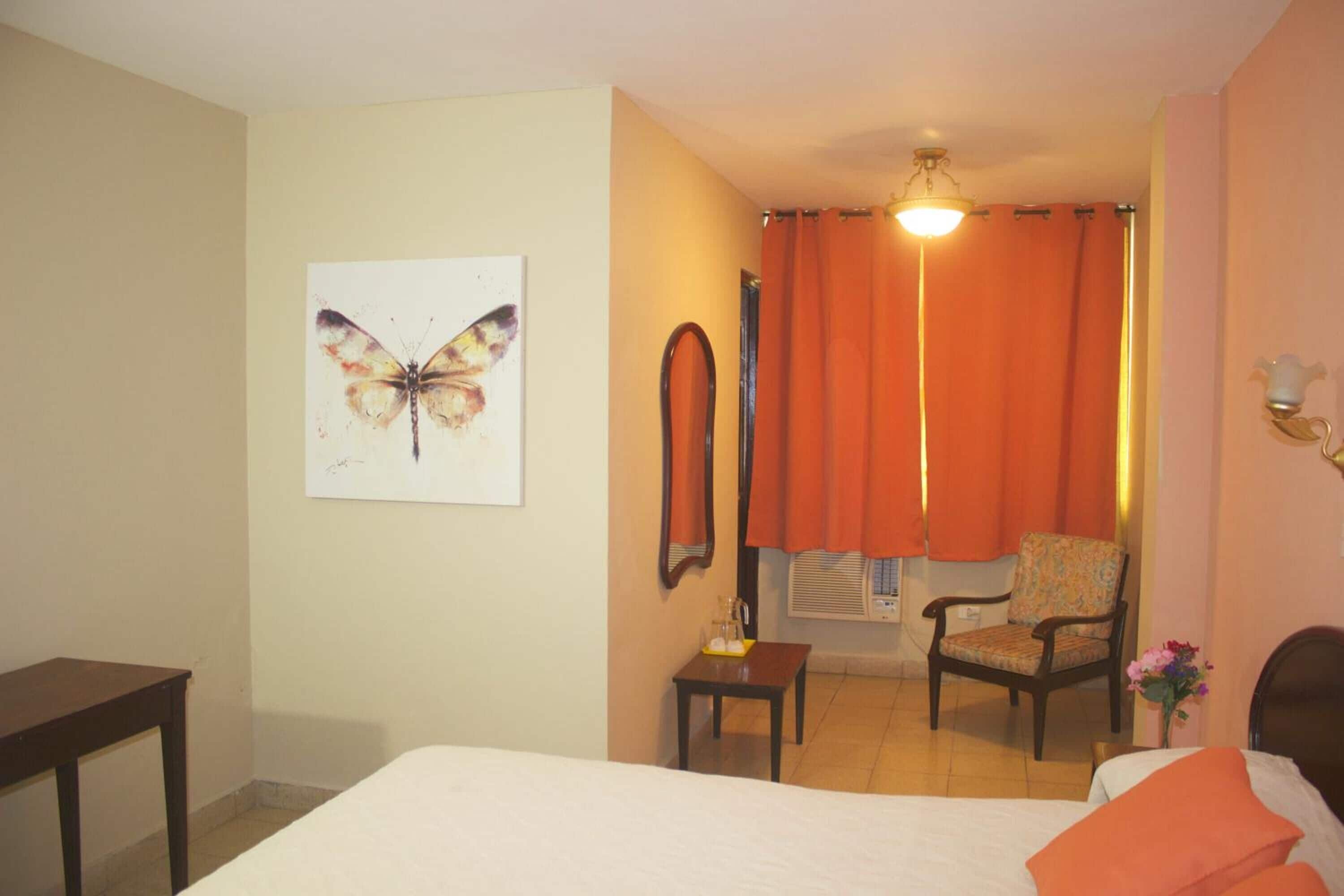 A hotel room with a bed, a chair, a small table, a mirror, and a butterfly artwork on the wall.