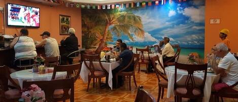 Un restaurante con mesas y sillas redondas, un mural de una playa y banderas en la pared.