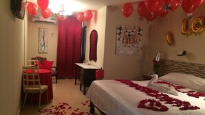 Habitación de hotel con una cama decorada con pétalos de rosa, un escritorio, una silla y globos rojos.
