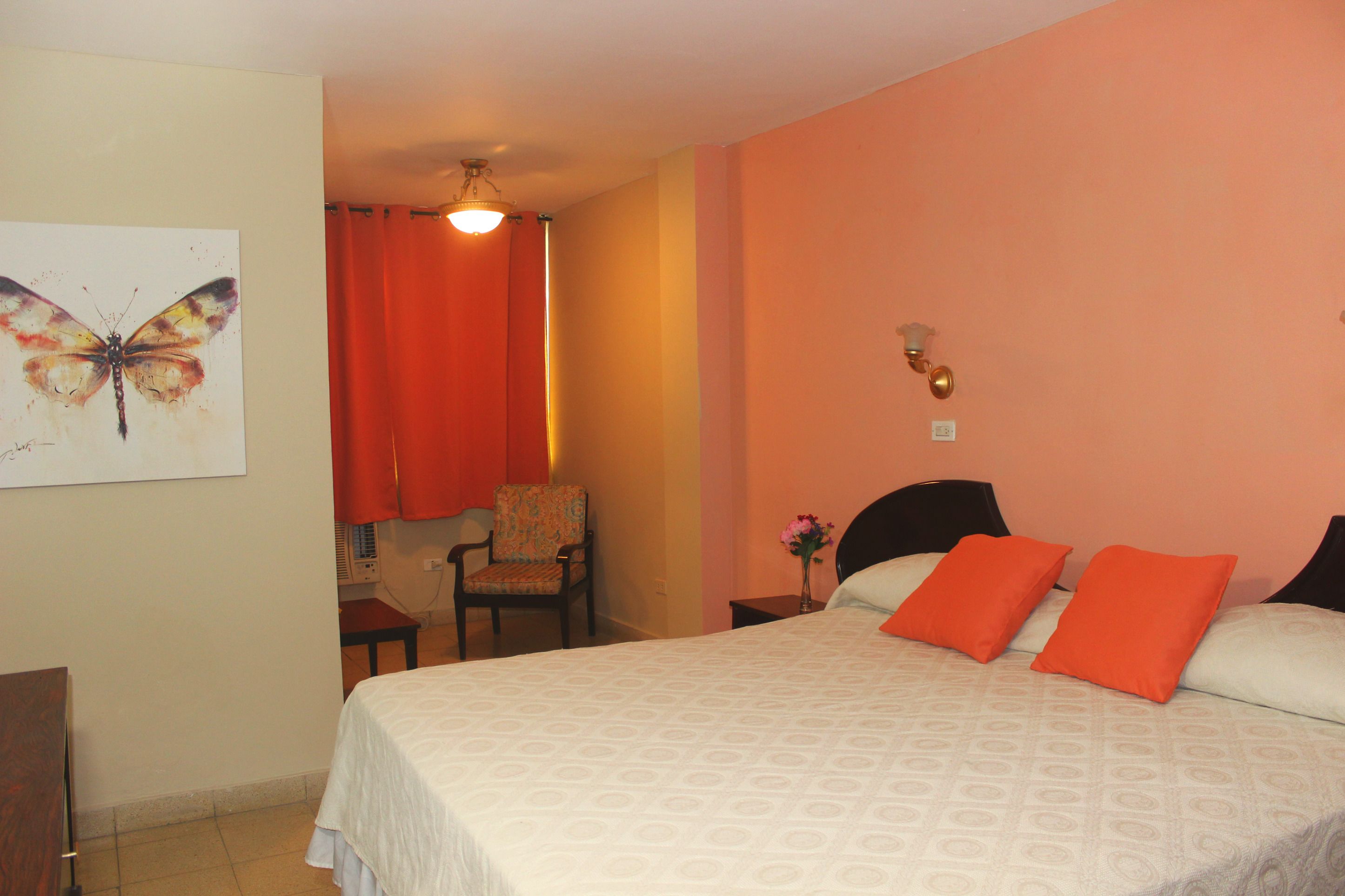 Un dormitorio con cama, una silla, una mesita de noche, una lámpara y un cuadro de mariposa en la pared.
