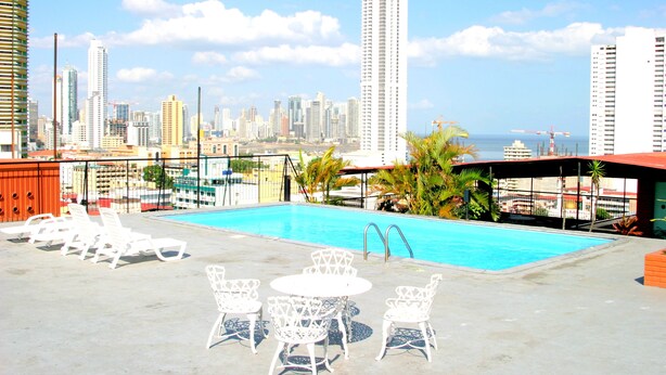 Zona de piscina en la azotea con sillones reclinables y una mesa, con vistas a la línea costera de la ciudad y al océano.
