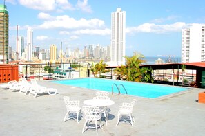 Zona de piscina en la azotea con sillones reclinables y una mesa, con vistas a la línea costera de la ciudad y al océano.