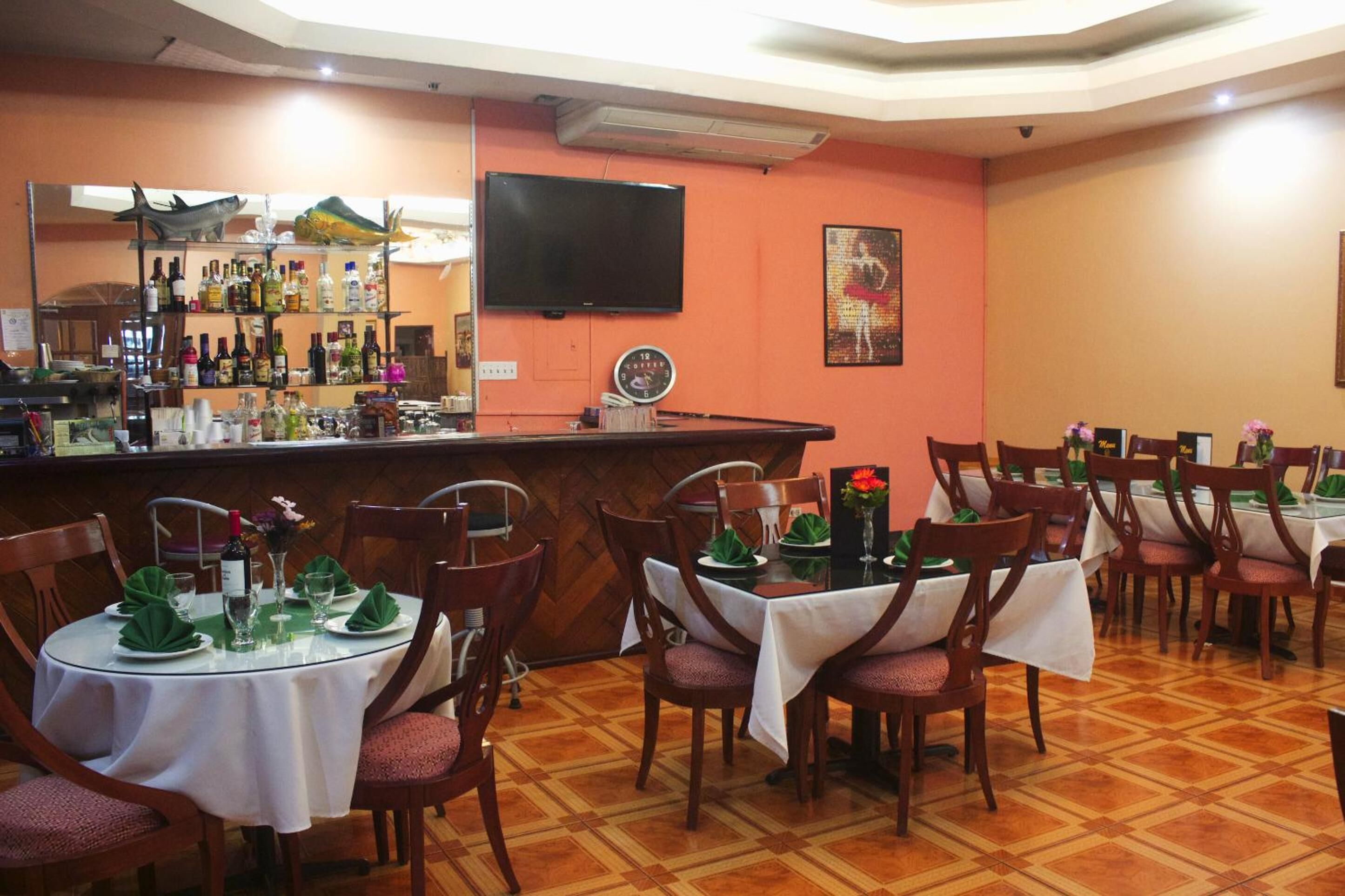 Un restaurante con varias mesas dispuestas para comer, una zona de bar con diferentes botellas y un televisor colgado en la pared.