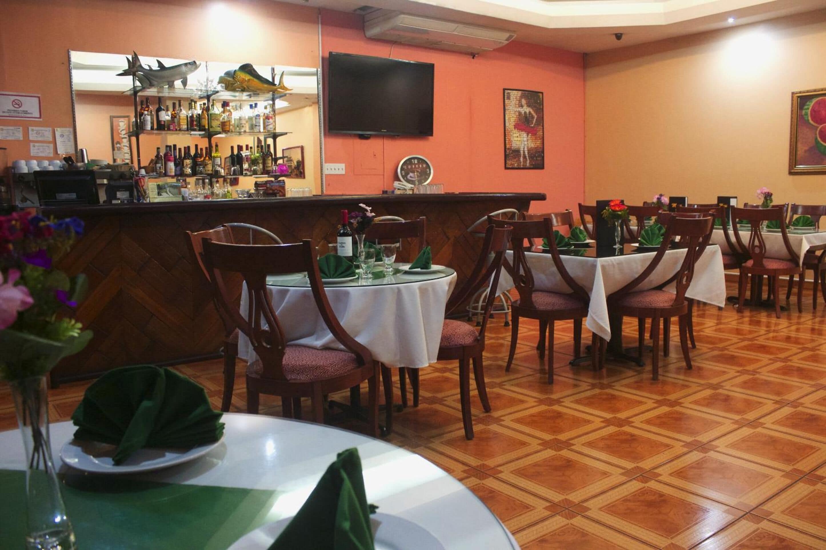 Un restaurant doté de nombreuses tables, de chaises, d’un bar avec des étagères remplies de bouteilles et d’un téléviseur fixé au mur.