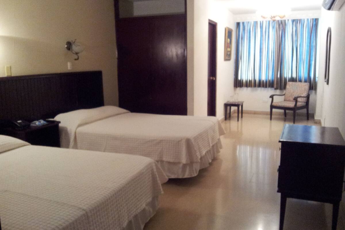 Habitación de hotel con dos camas, un escritorio, una silla, una mesita y una puerta.