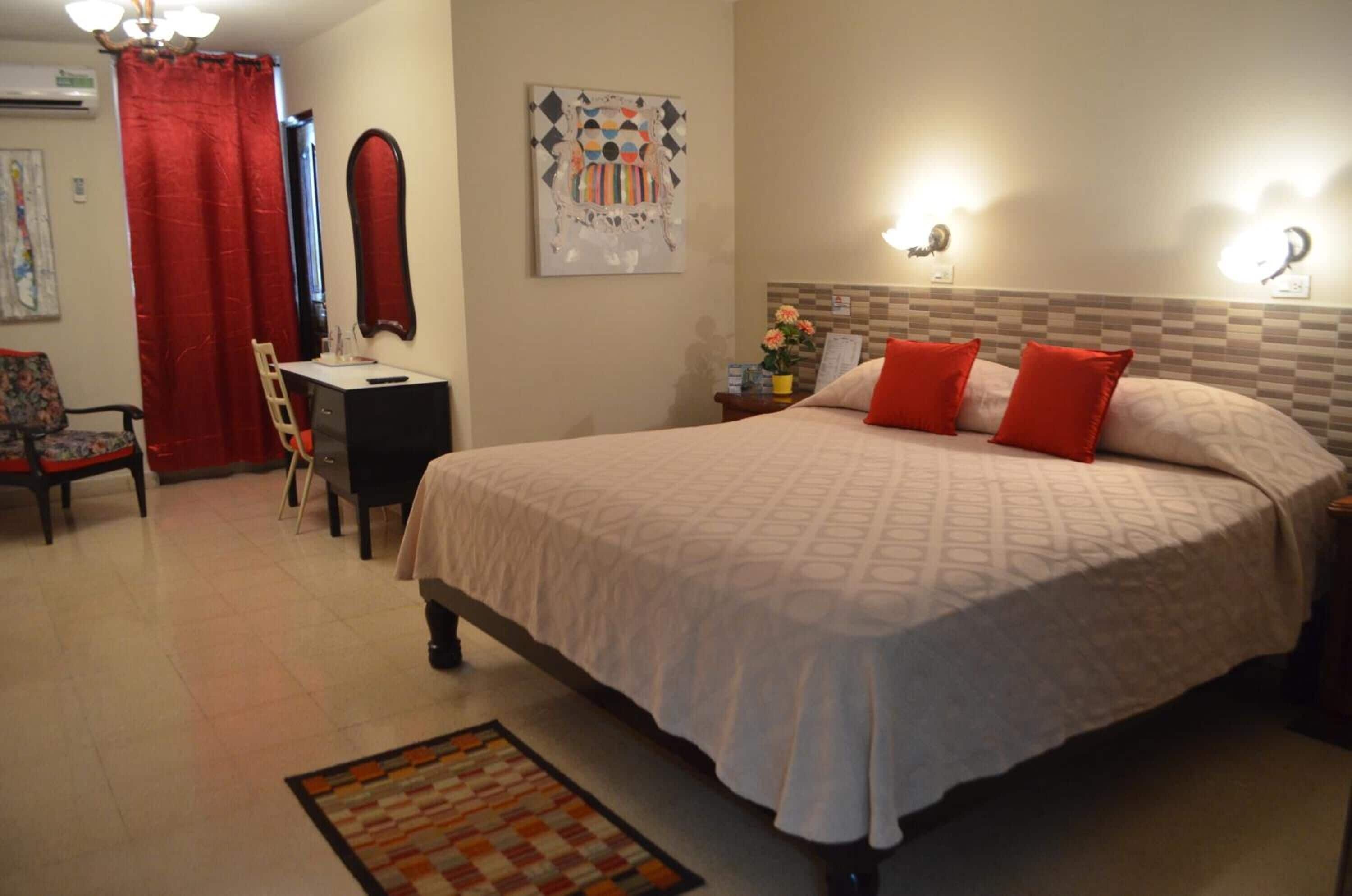 Habitación de hotel con cama, escritorio, silla, cortina roja y una obra de arte colgada en la pared.