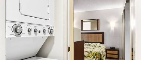 A laundry room with a washer and dryer, a wooden cabinet, a bed with a tropical print, and a mirror on the wall.
