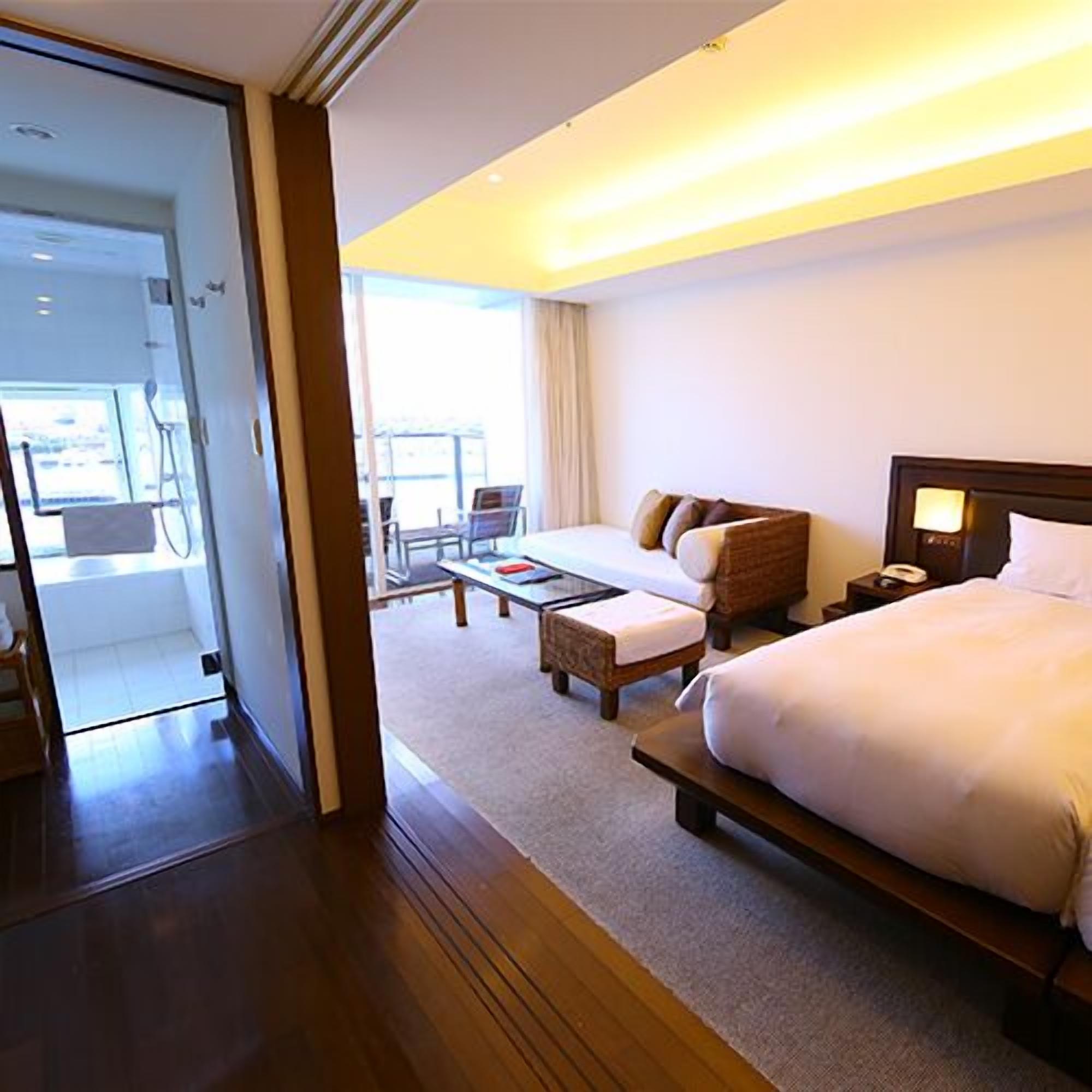 Quarto de hotel com uma cama grande, um sofá, uma mesinha e uma cadeira. Uma varanda é visível pela porta.