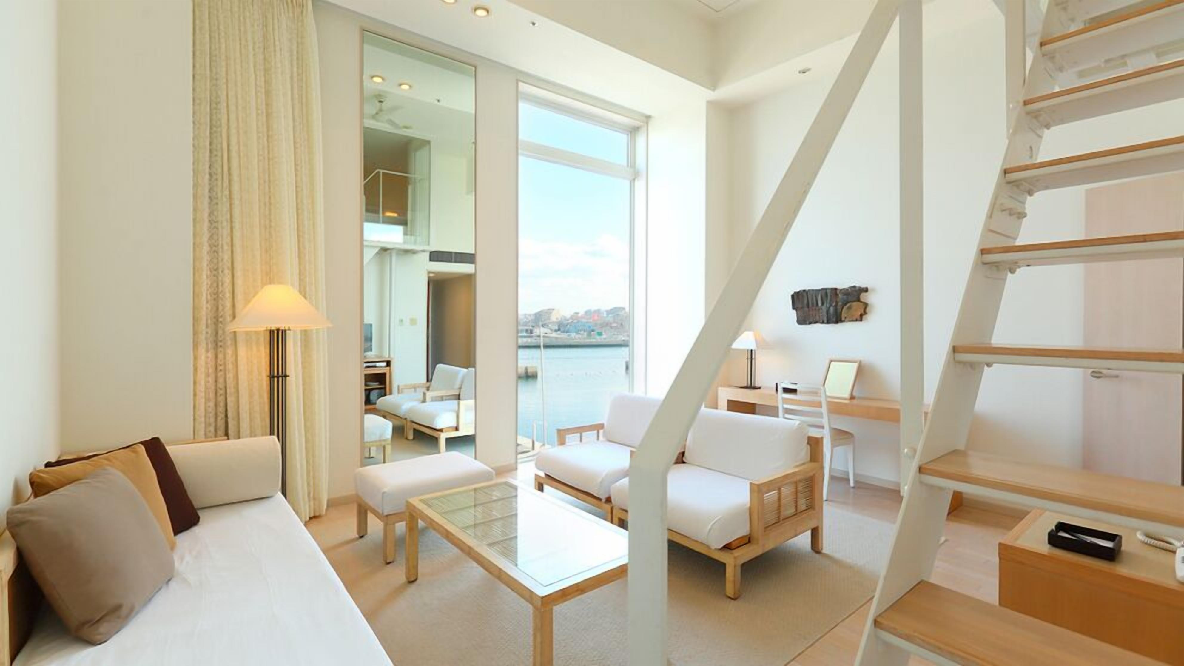 Quarto de hotel moderno com uma janela grande, um sofá, uma mesa de centro e uma escrivaninha com uma cadeira.