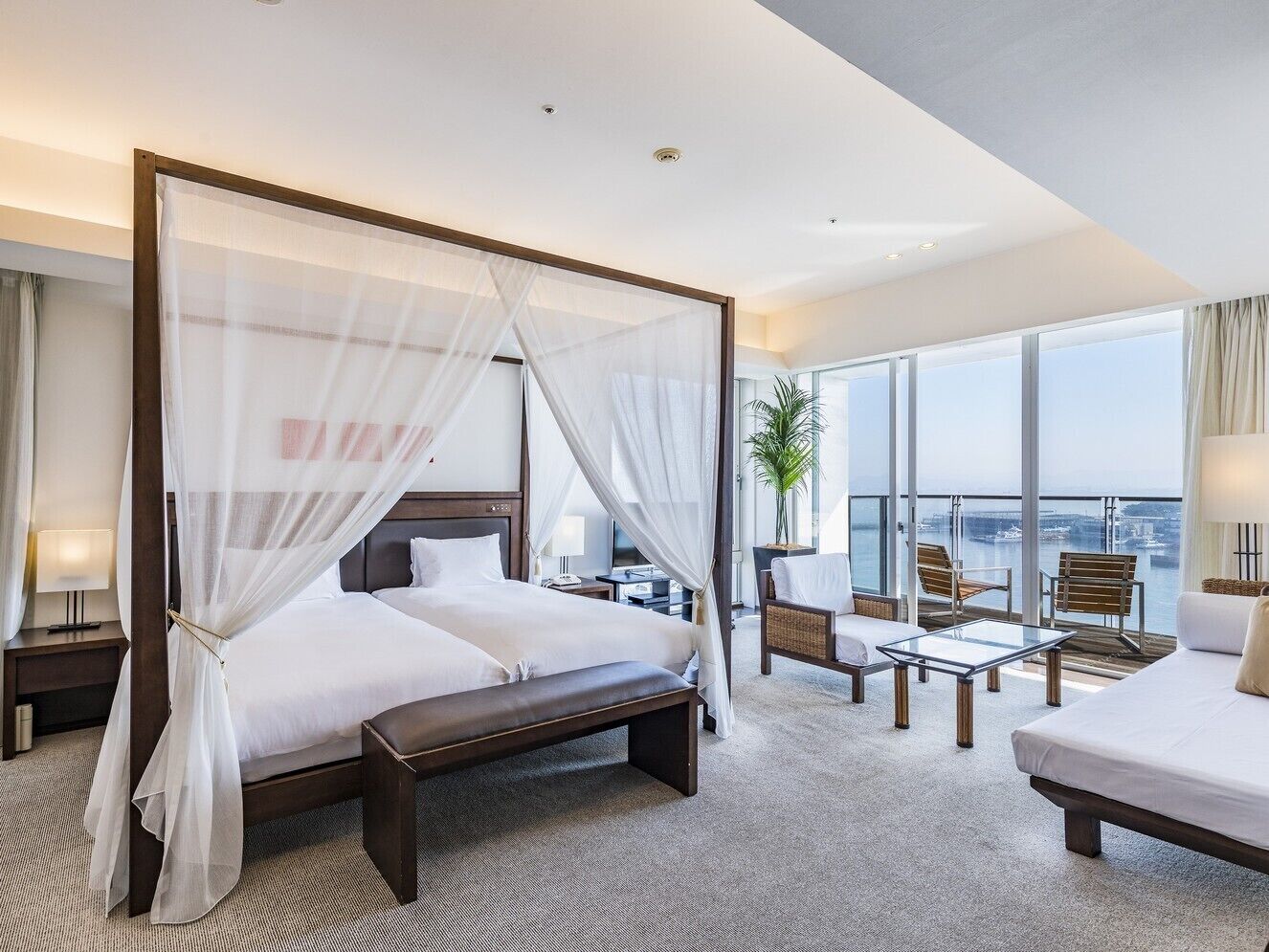 Quarto de hotel moderno com cama de dossel, varanda com assentos e uma grande janela com vista para a cidade.