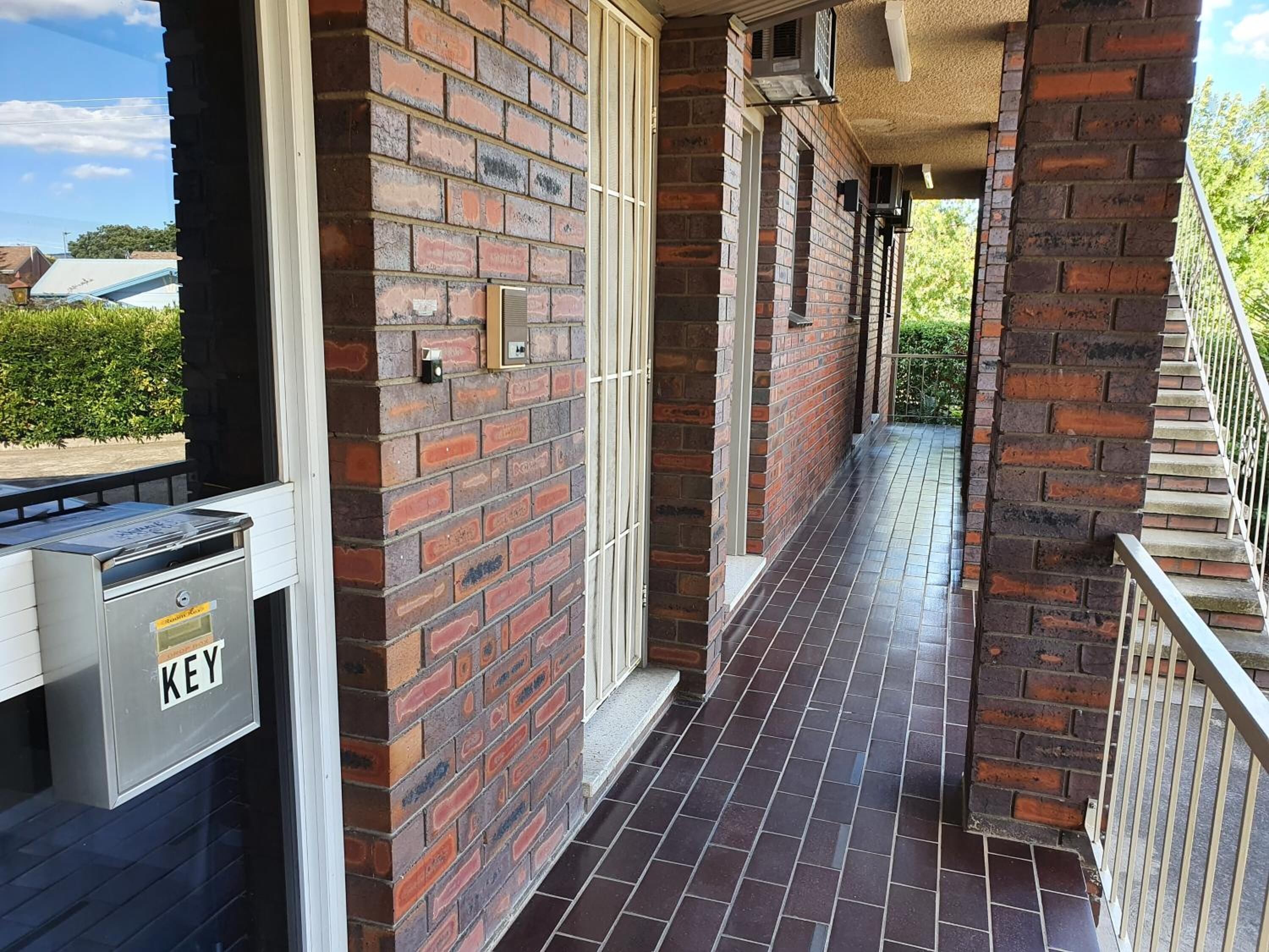 Murstensfacade med et metalpostkasse med teksten "KEY" og en fliseterrasse.