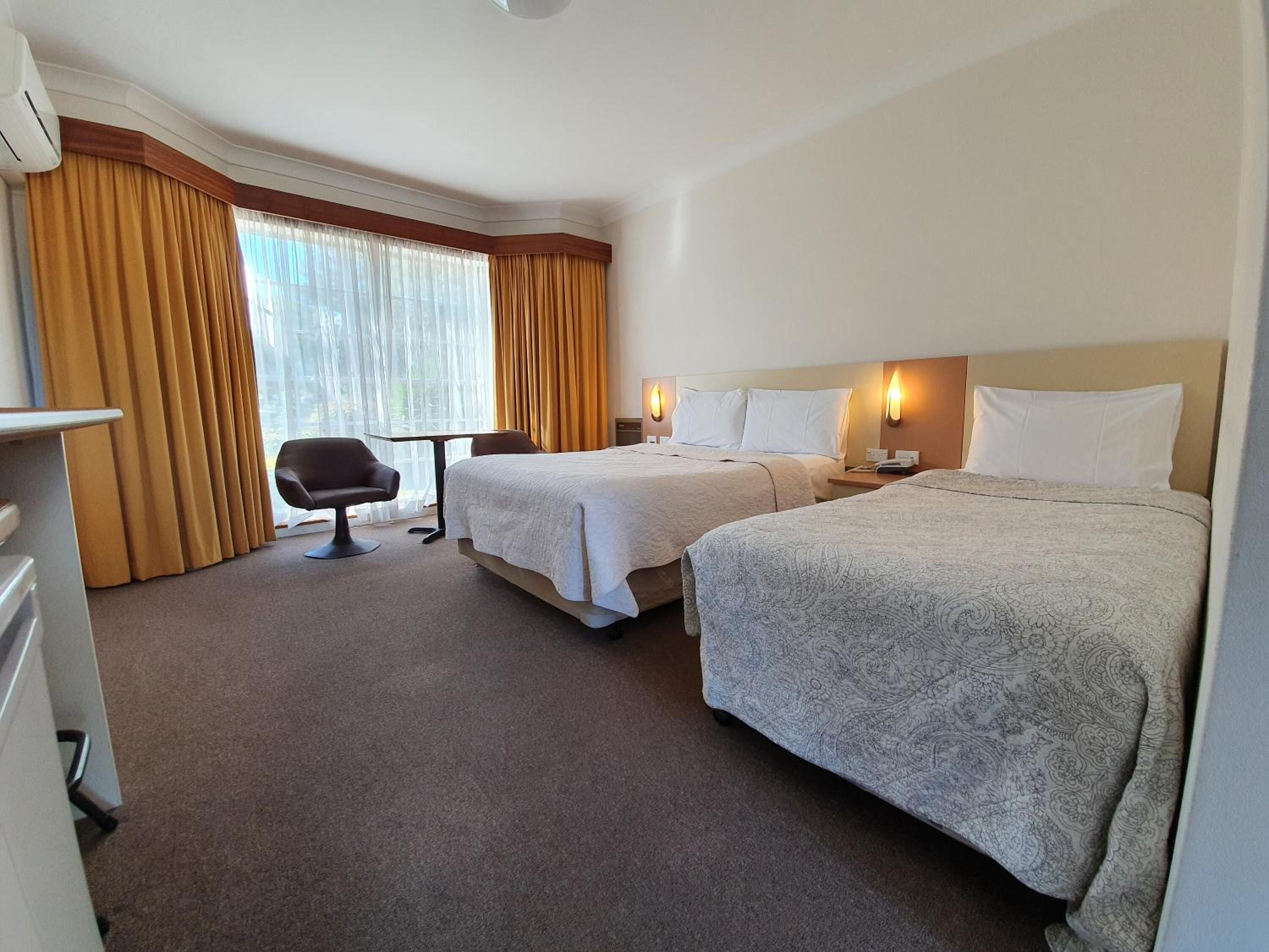 Habitación de hotel con dos camas, un escritorio, una silla y un ventanal con cortinas.