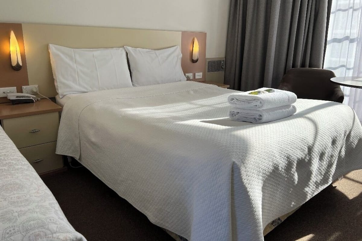 A neatly made bed with white linens, a bedside table with a lamp, and a chair by the window.