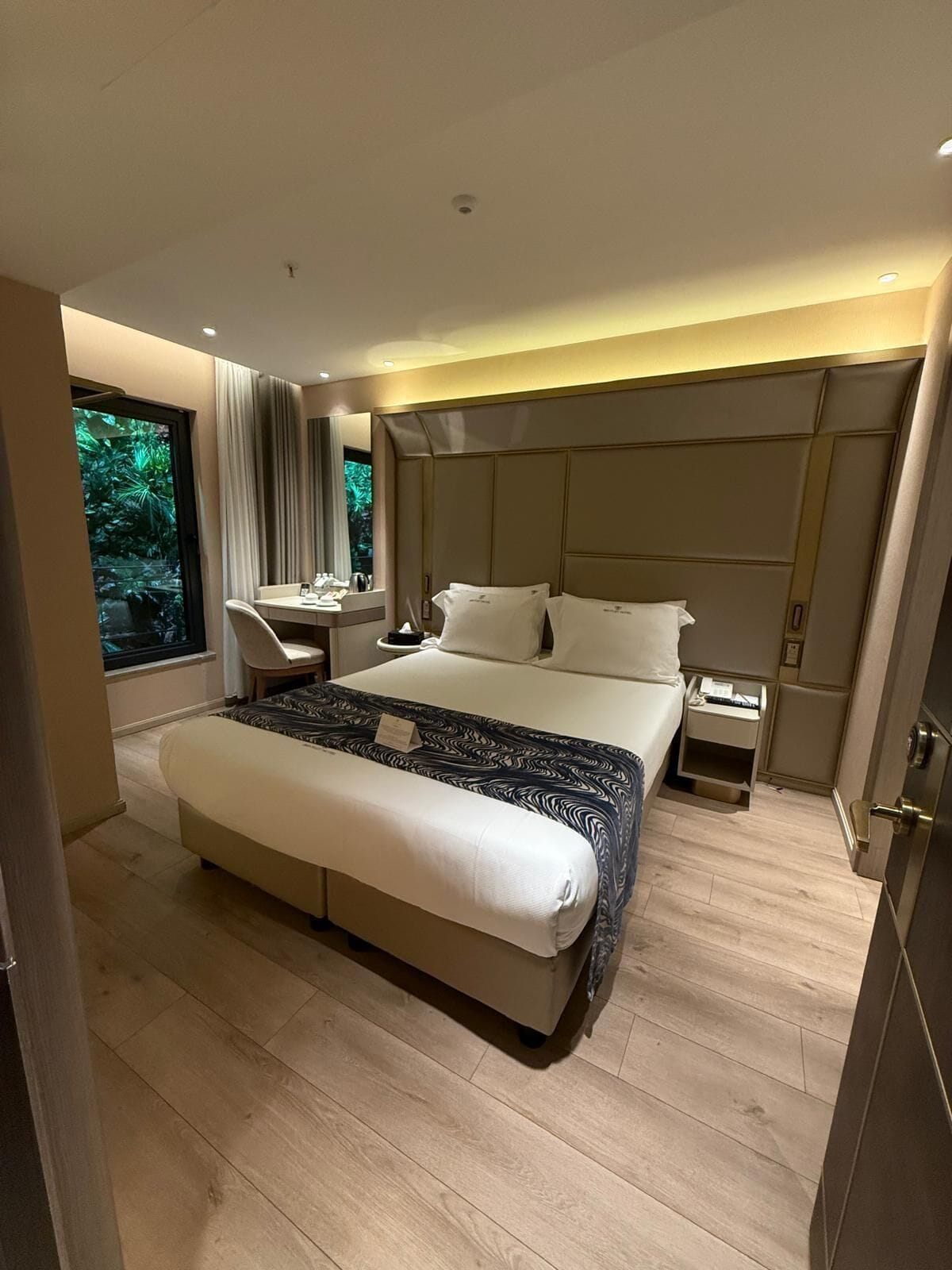 A modern hotel room with a large bed, a desk, a nightstand, and a view of greenery through a window.