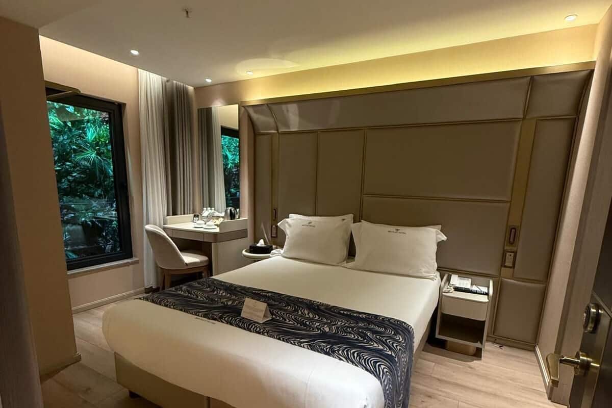 A modern hotel room with a large bed, a desk, a nightstand, and a view of greenery through a window.