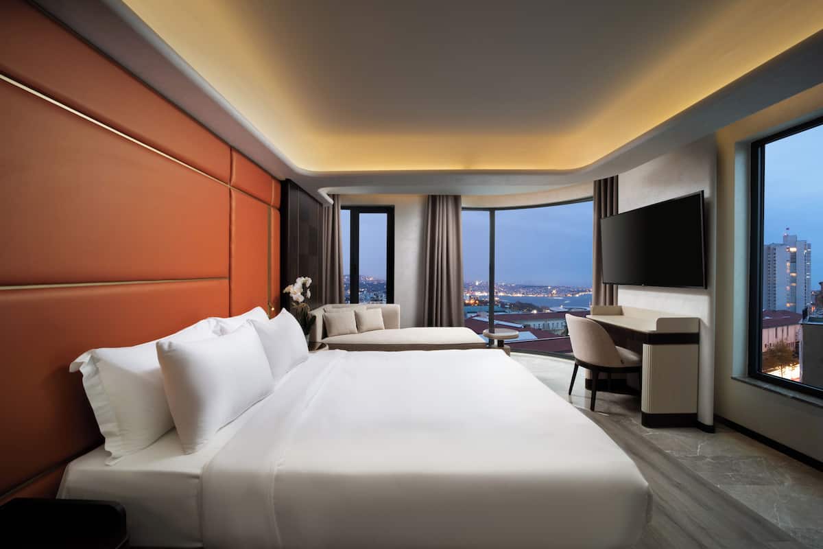 A hotel room with a large bed, a desk, a chair, and a TV. There is a view of a cityscape through large windows.