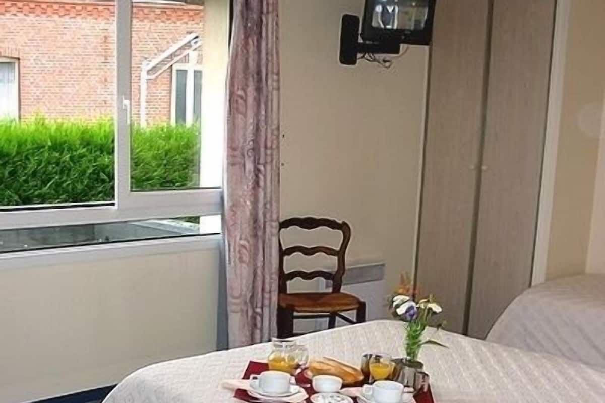 Quarto de hotel com uma cama, uma janela com vista para vegetação e um edifício de tijolos, e um tabuleiro de café da manhã sobre a cama.