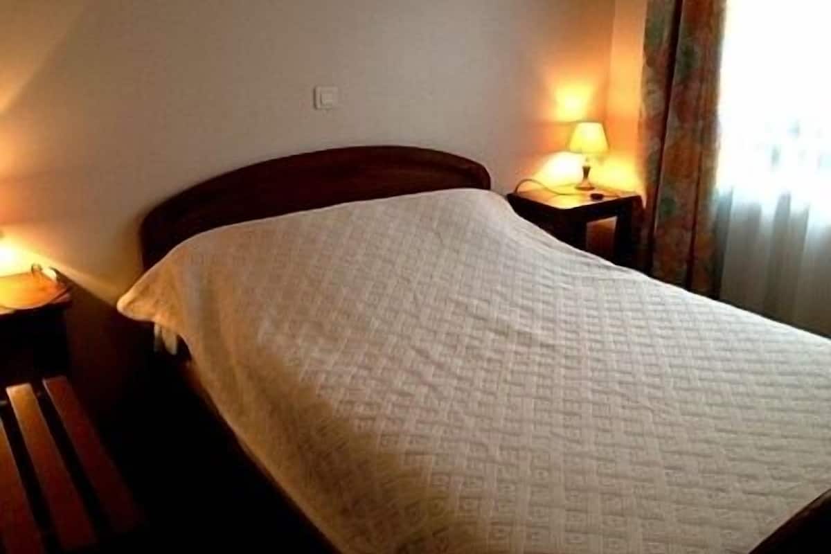 Uma cama bem arrumada com colcha branca, duas luminárias de apoio e cabeceira de madeira.