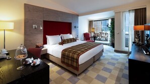 Een hotelkamer met een groot bed, een bureau met lamp, een zithoek met tafel en stoelen, en uitzicht op de stad door het raam.