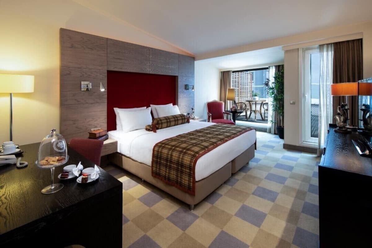 A hotel room with a large bed, a desk with a lamp, a dining area with a table and chairs, and a view of the city through the window.