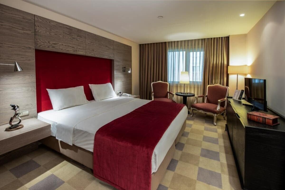 A hotel room with a large bed, two armchairs, a desk with a TV, and a checkered floor.