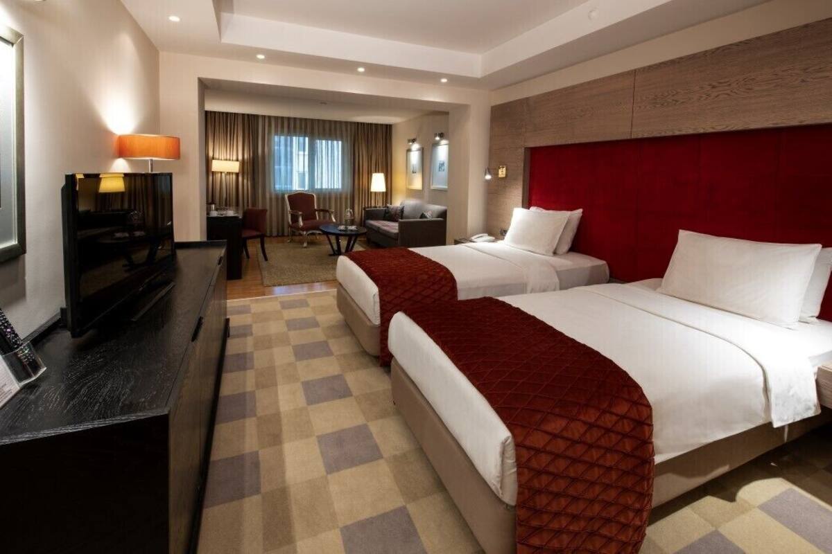 A hotel room with two beds, a desk, a TV, and a seating area with a chair and table.