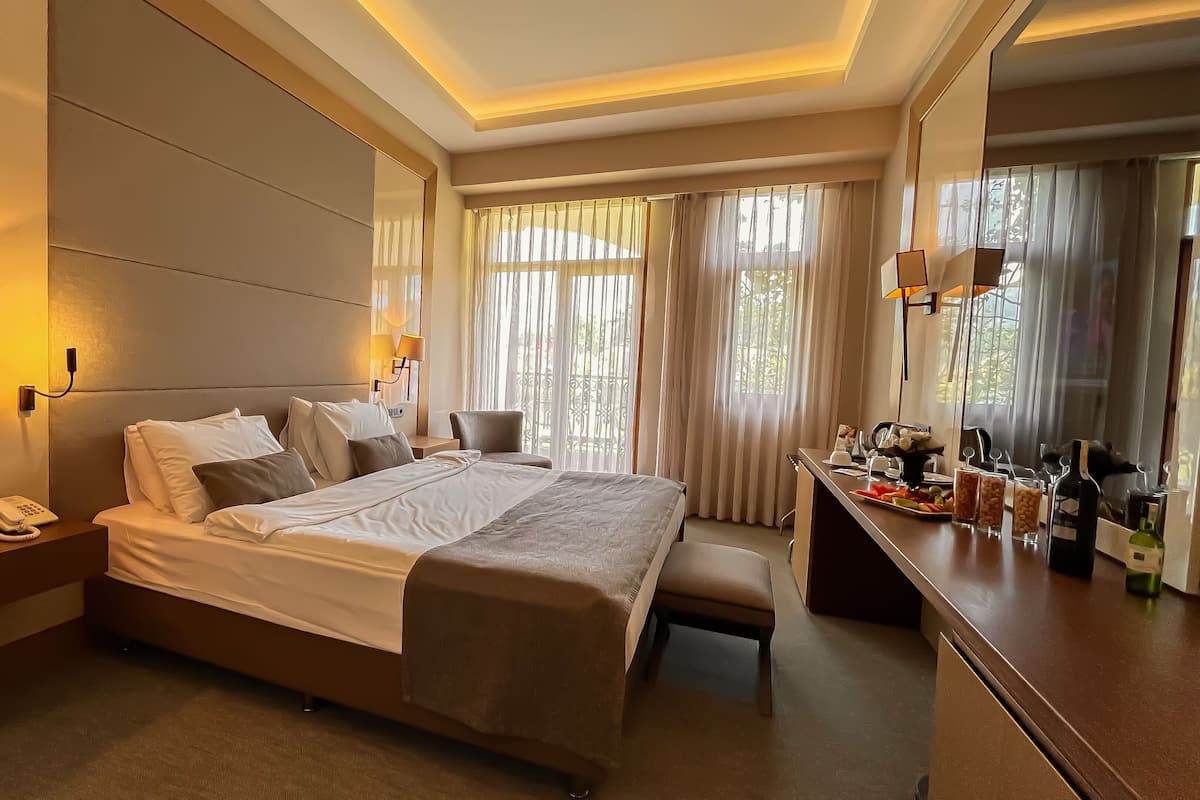 A hotel room with a large bed, bedside lamps, a desk with a bottle of wine, and a view of greenery through the window.
