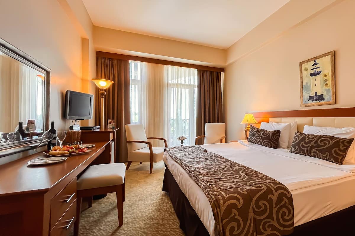 A hotel room with a large bed, a desk with a computer, a chair, a TV, and a painting on the wall.