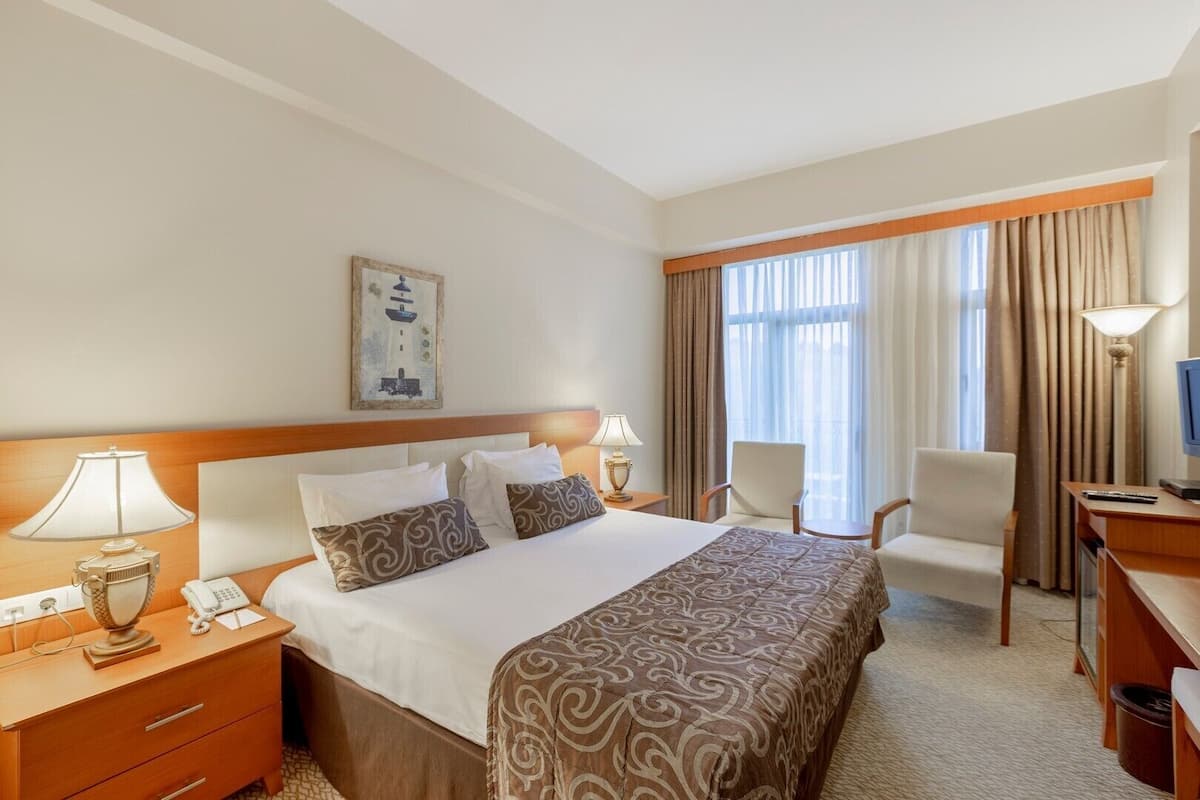 A hotel room with a large bed, two bedside tables, a desk with a chair, a television, and a window with curtains.