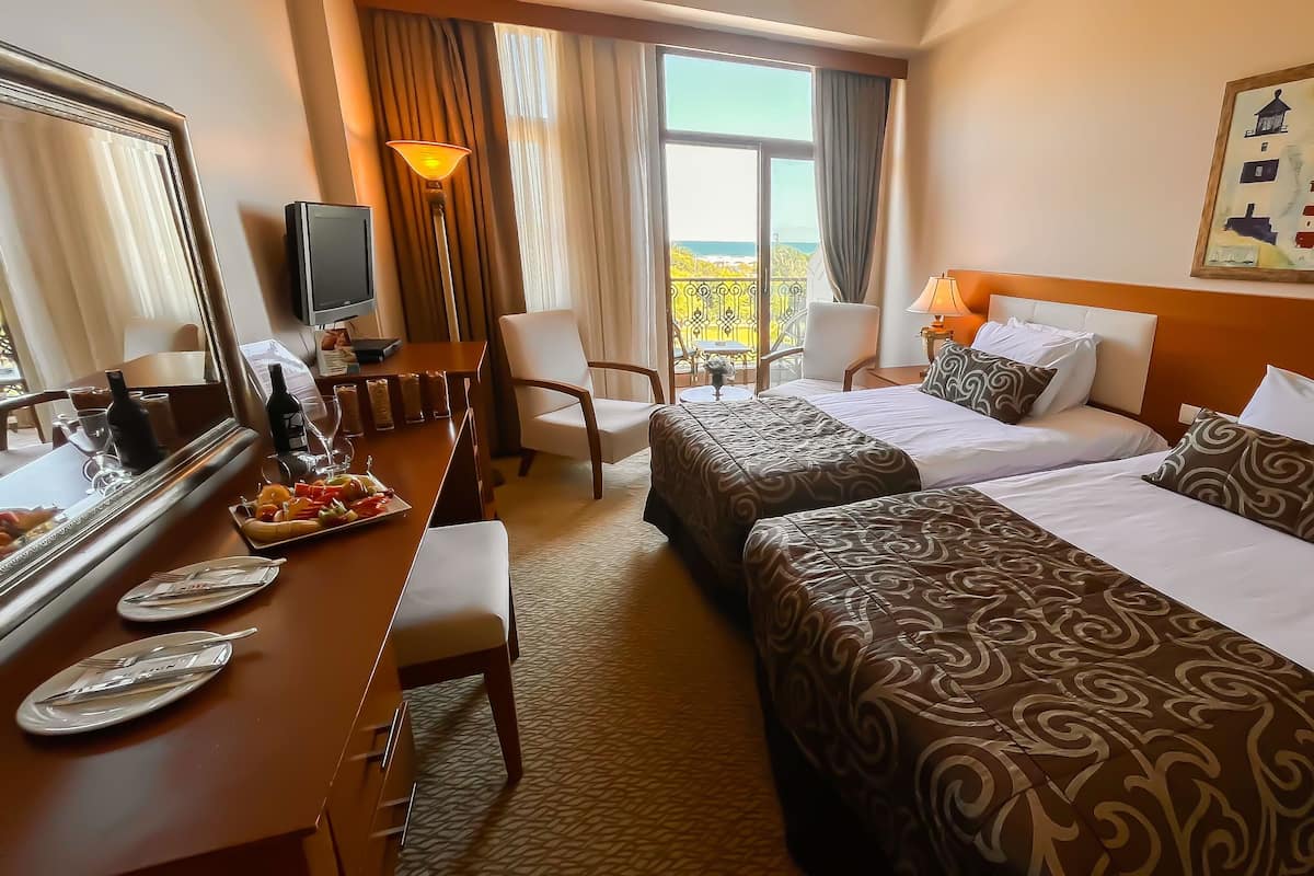 A hotel room with two beds, a desk with a mirror, a dining table with plates and cutlery, and a view of the outdoors.