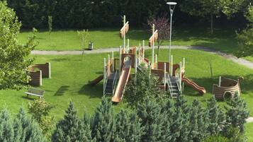Children can play on the slides, climbing structures, and a boat-shaped play area surrounded by lush grass and trees.