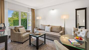 Un salon moderne avec un canapé, un fauteuil et une table basse en verre. On y trouve une grande fenêtre avec des rideaux, un lampadaire et un miroir.