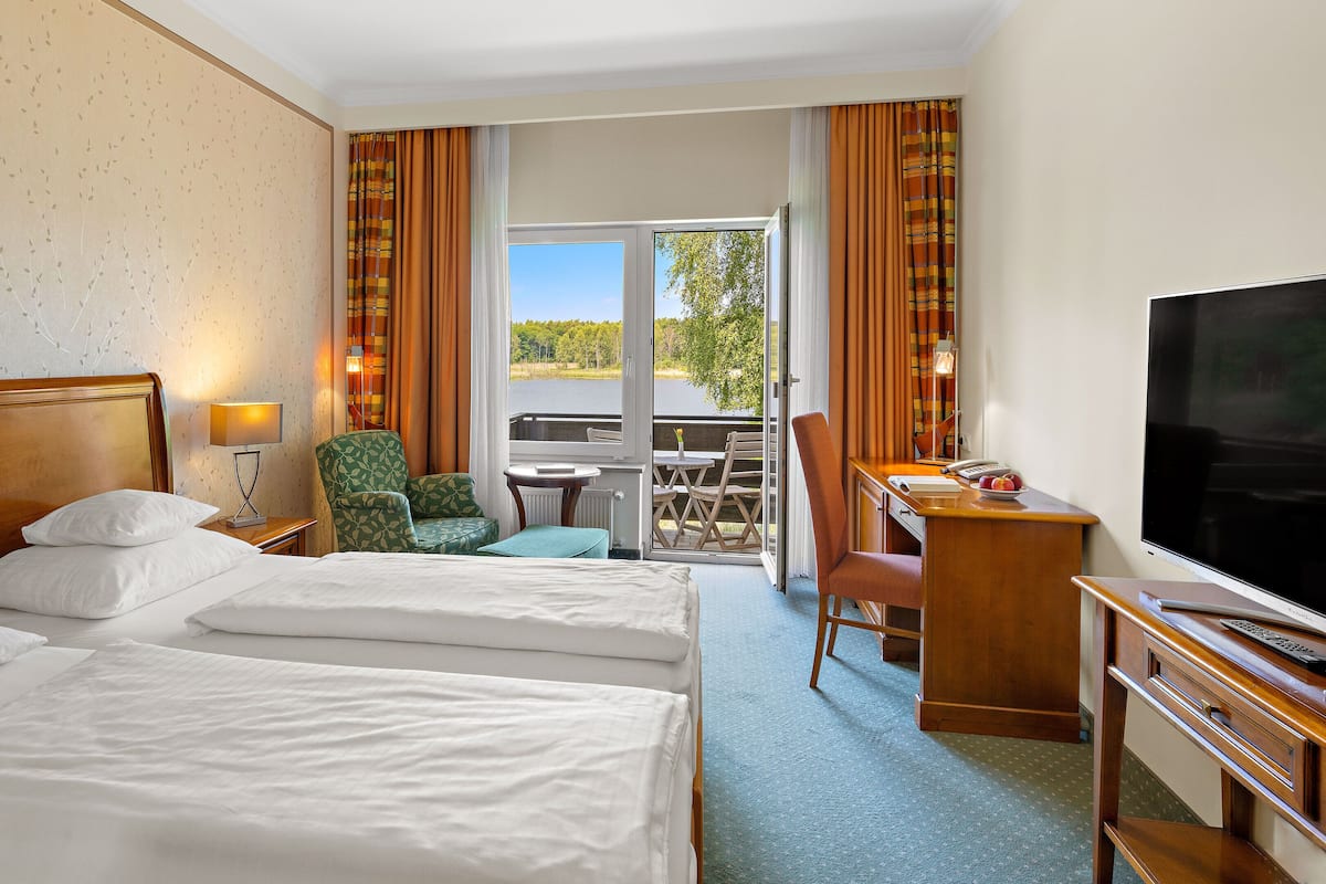 Quarto de hotel com duas camas, uma mesa com televisão, uma cadeira, uma mesinha e vista para um lago.
