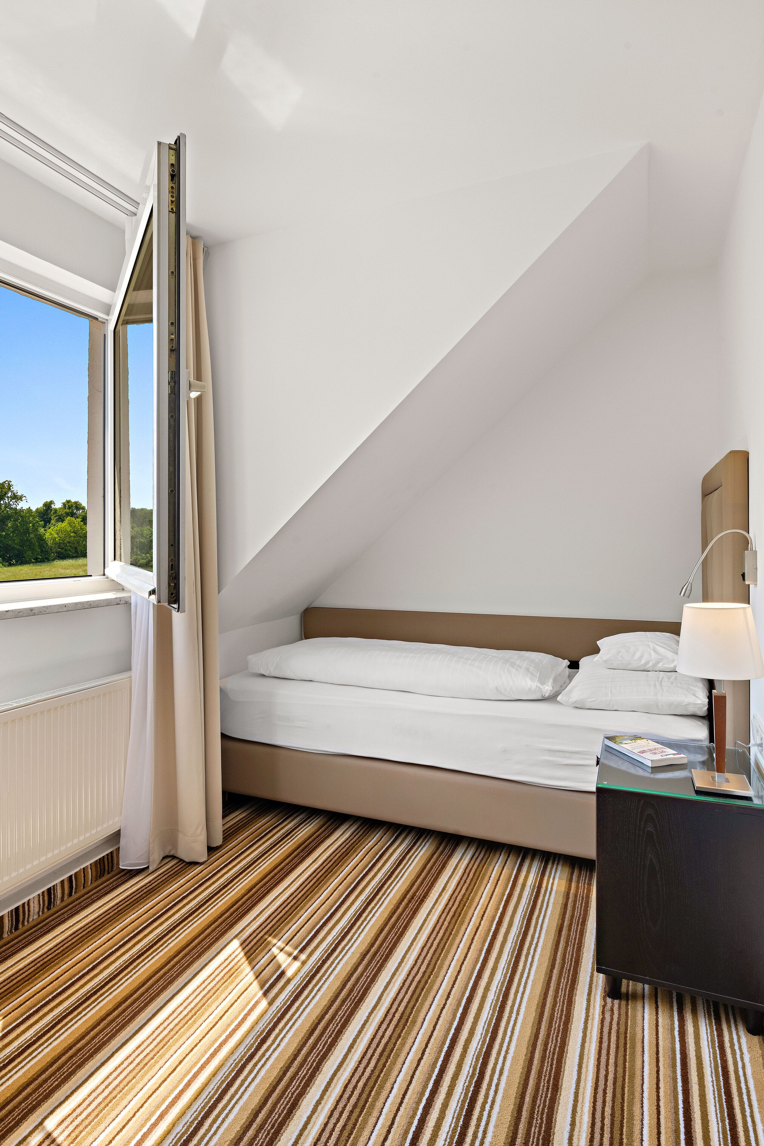Un dormitorio moderno con techo inclinado, una cama con ropa de cama blanca, un cabecero de madera, una mesita de noche con una lámpara y vistas a la vegetación a través de una ventana.