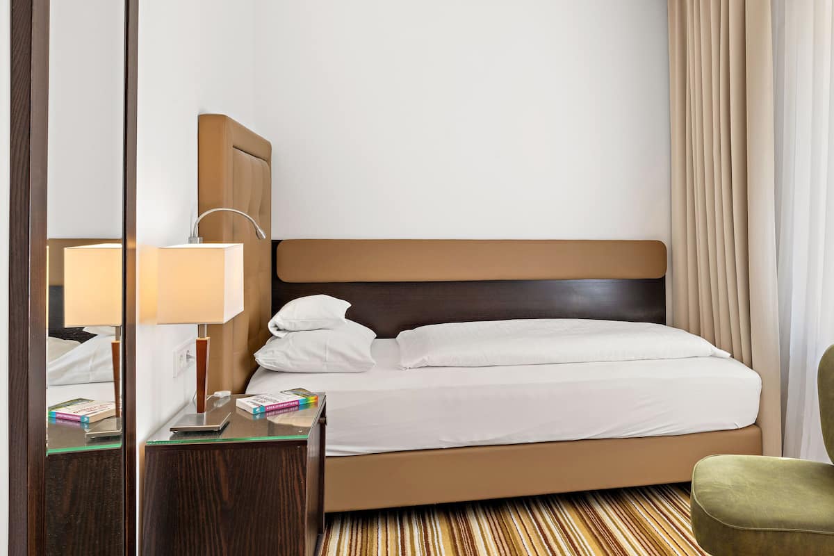 Cama bem arrumada com lençóis brancos, cabeceira de madeira, mesa de cabeceira com abajur e poltrona verde.
