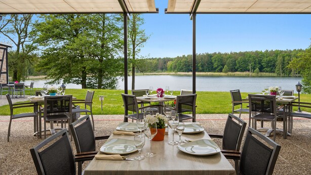 An outdoor dining area with tables set for two, overlooking a lake and surrounded by trees. - Seehotel Mühlenhaus (Chorin)
