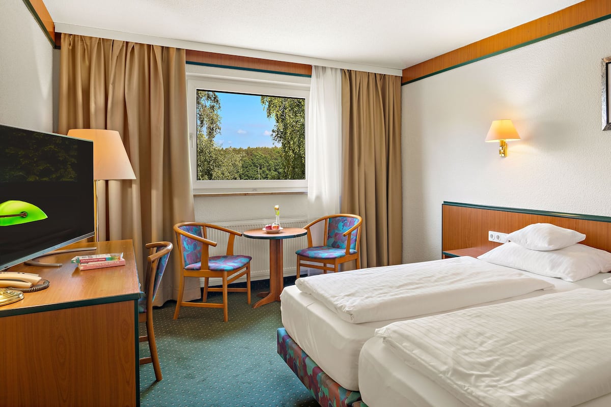Quarto de hotel com duas camas, uma escrivaninha com televisão, uma cadeira, uma mesinha e uma janela com cortinas.