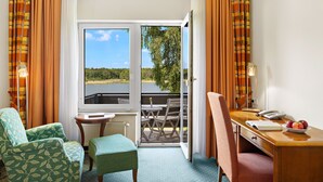 A hotel room with a balcony, a desk with a telephone, two chairs, a small table, and a view of a lake and trees. - Seehotel Mühlenhaus (Chorin)