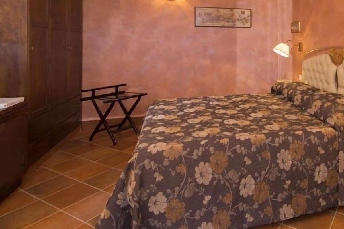A bedroom with a floral bedspread, a wooden wardrobe, a bedside table with a lamp, and a wall-mounted phone.