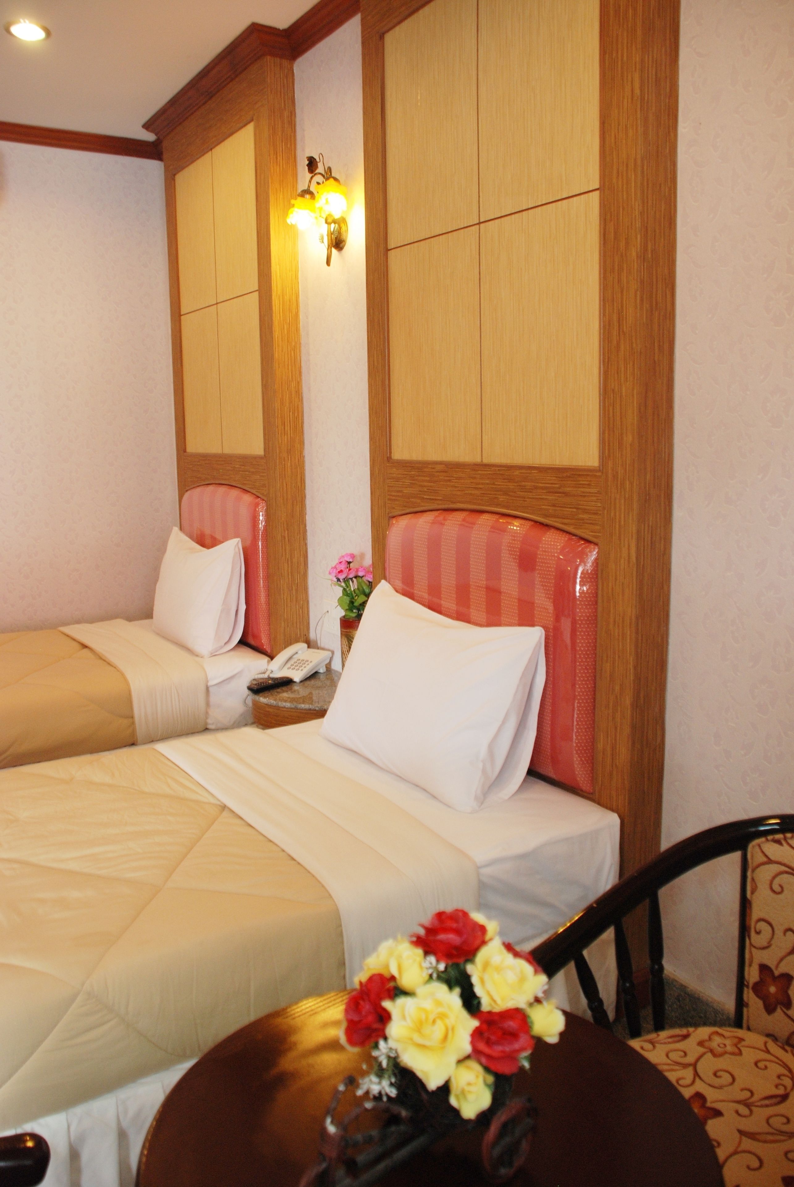 A hotel room with two beds, a wooden headboard, a bedside table with a phone, and a vase with flowers.