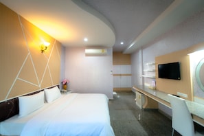A hotel room with a bed, bedside table, lamp, air conditioning unit, desk, chair, and a flat-screen TV mounted on the wall.