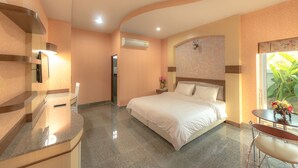 A hotel room with a bed, a desk, a chair, a nightstand, and a window with a view of greenery. - Martina Hotel (Surin)