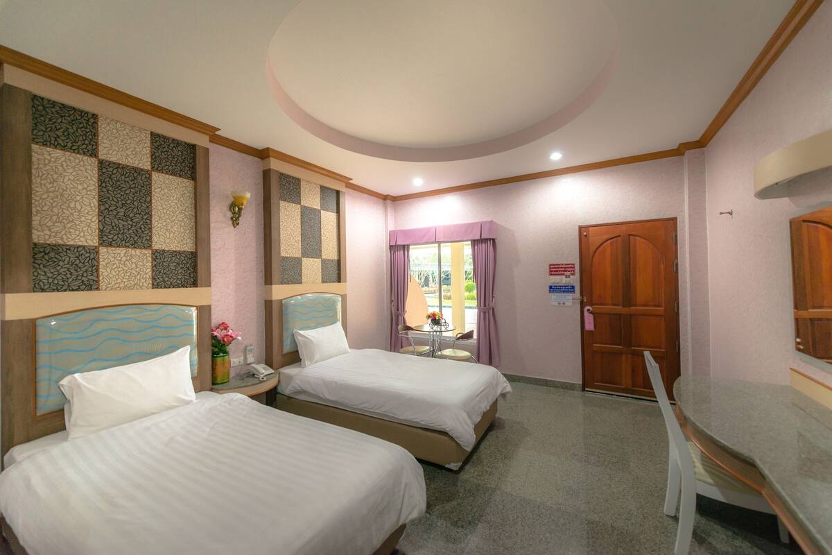 A hotel room with two beds, a small table with a chair, a wall-mounted lamp, and a door with a sign.