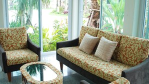 A room with a sofa and two armchairs, a glass coffee table, and large windows offering a view of palm trees and a garden. - Martina Hotel (Surin)