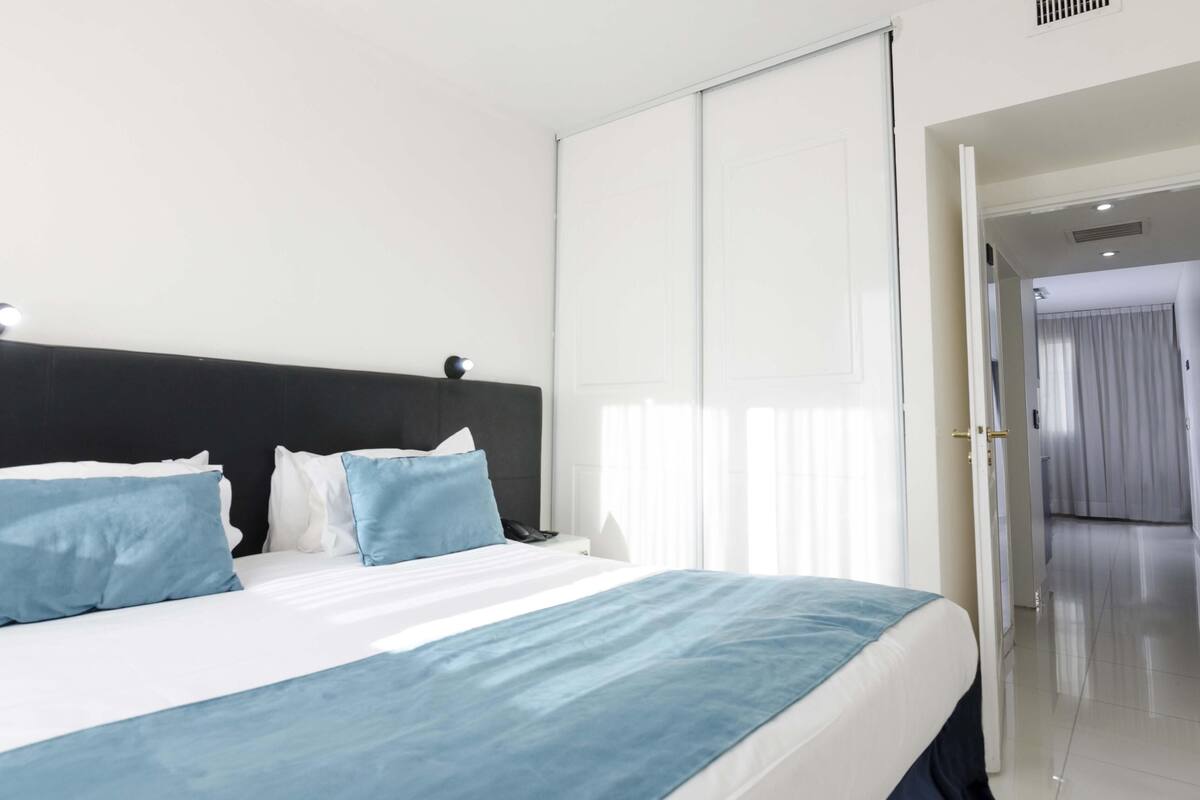 A hotel room with a bed, a nightstand, a window with curtains, and a door leading to another room.