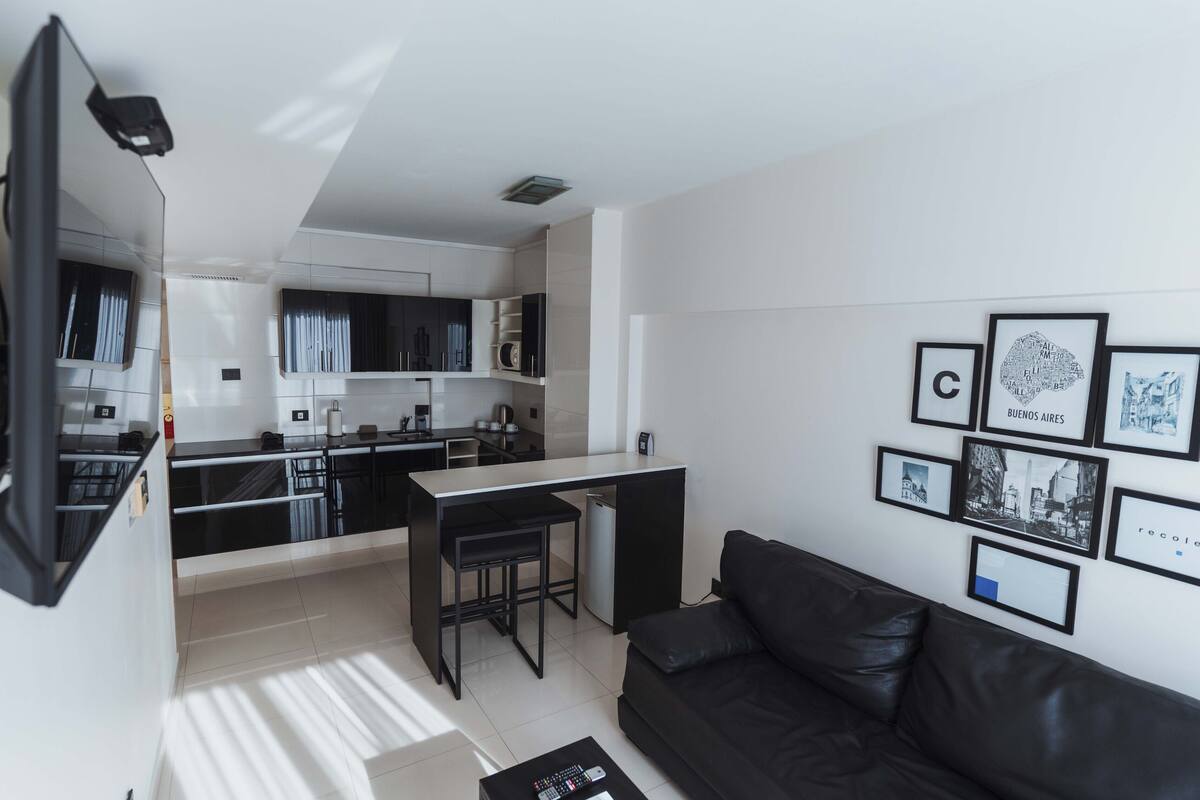 A modern kitchen with a dining area, a black leather sofa, and framed pictures on the wall.
