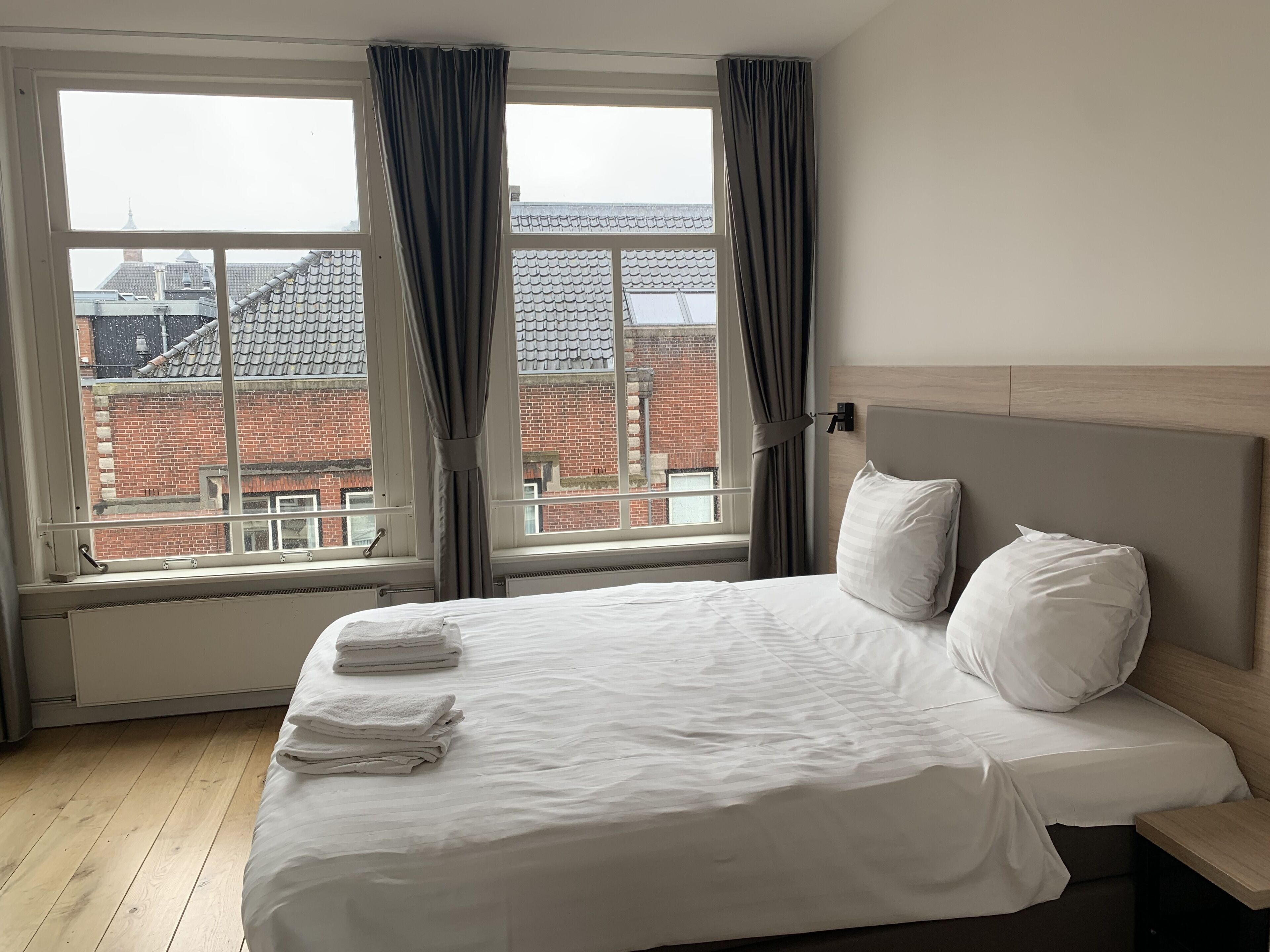 Een hotelkamer met een groot bed, twee kussens, een houten hoofdbord en een raam met gordijnen.