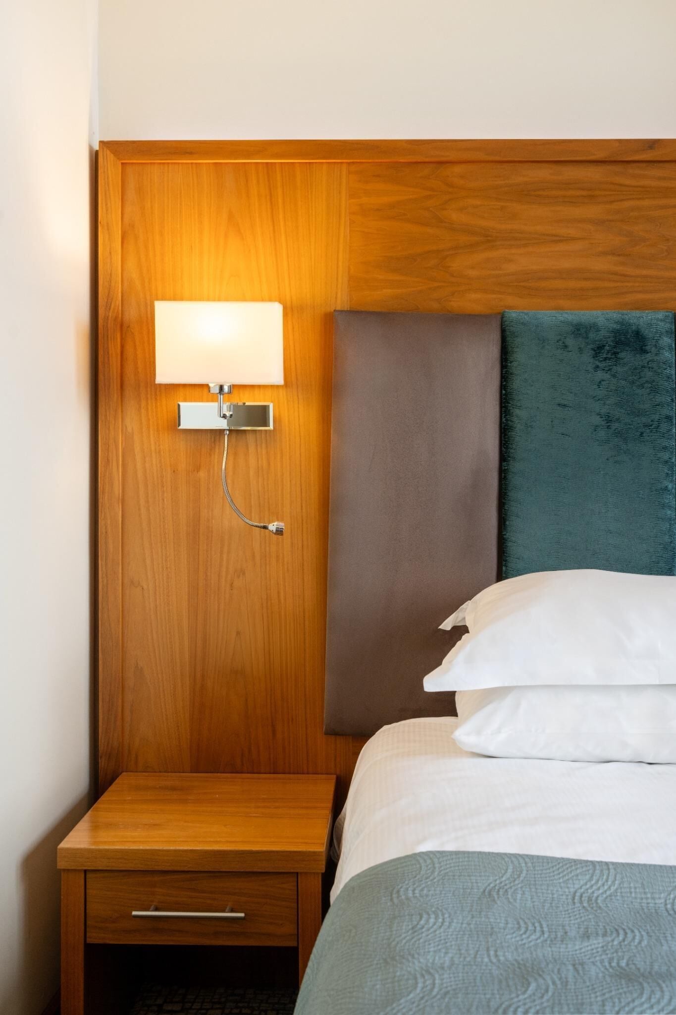 Een hotelkamer met een houten hoofdbord, een bedlampje, een nachtkastje met lade, en een bed met wit en teal beddengoed.